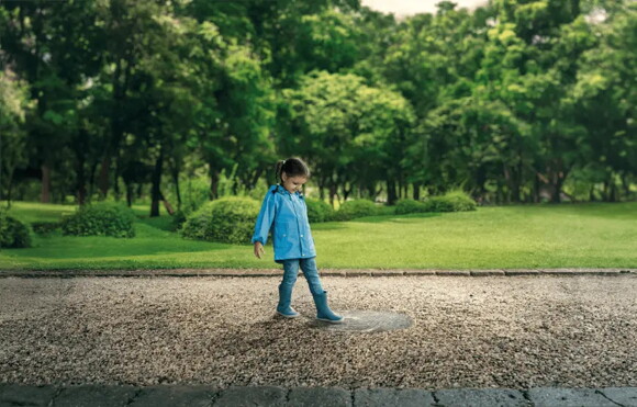 Niña con impermeable azul y botas de lluvia, mirando un charco en un camino de grava en un parque verde y frondoso